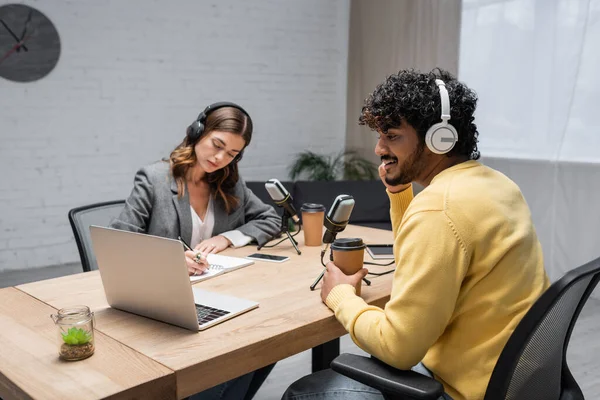 Cheerful and curly indian man in headphones and yellow jumper holding coffee to go and looking at laptop near microphones and radio host writing in notebook near smartphone on table in radio studio — Stock Photo