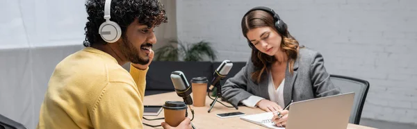 Curly and happy indian man in headphones and yellow jumper sitting with paper cup near laptop and radio host writing in notebook close to smartphone with blank screen in broadcasting studio, banner — Stock Photo