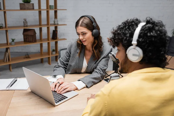 Smiling brunette broadcaster in grey blazer typing on laptop near microphones and notebook while blurred indian man in yellow jumper sitting in headphones in radio studio — Stock Photo