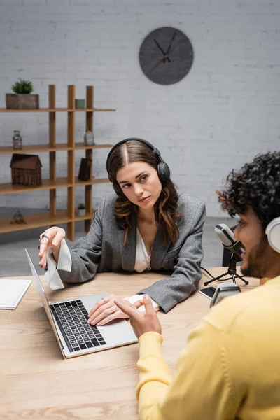 Brunette broadcaster in headphones and grey blazer looking at curly indian man pointing at laptop near microphones and smartphone on table while recording interview in modern radio studio — Stock Photo