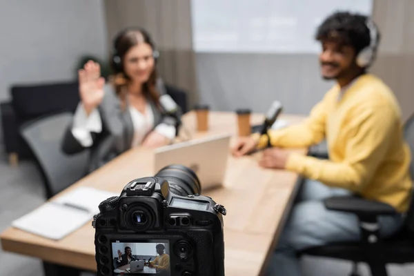 Entrevista profesional de grabación de cámara digital de mujer saludando de la mano y joven invitado indio sentado en la mesa con el ordenador portátil y tazas de papel borroso en el estudio de radiodifusión - foto de stock