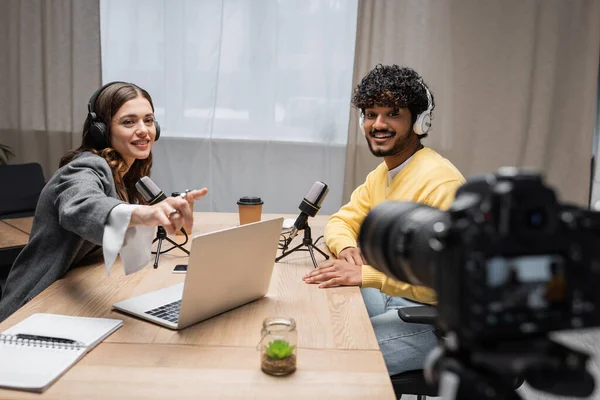 Cheerful broadcaster in headphones pointing at laptop near coffee to go and notebook during interview with curly indian guest in yellow jumper in front of blurred digital camera in modern radio studio — Stock Photo