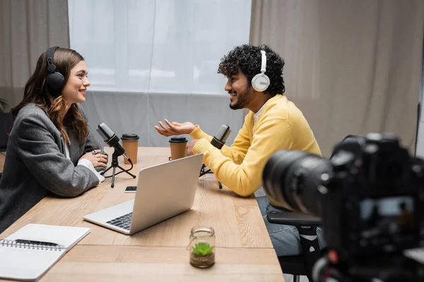 Side view of curly and smiling indian man in headphones and yellow jumper talking to brunette interviewer near laptop, notebook and takeaway drinks in front of blurred digital camera in radio studio — Stock Photo
