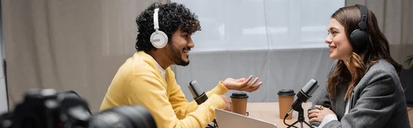 Vista lateral del hombre indio feliz con auriculares y jersey amarillo haciendo gestos y hablando con el entrevistador sonriente con chaqueta gris en el estudio de radio cerca de vasos de papel y micrófonos profesionales, pancarta - foto de stock