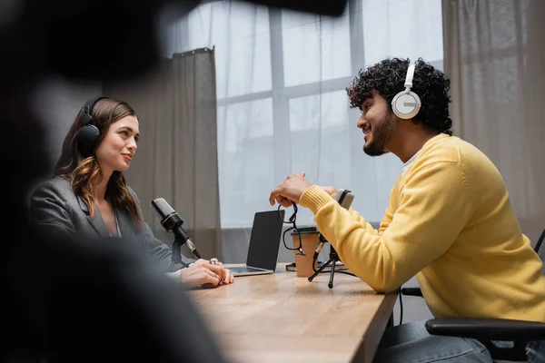 Side view of smiling and curly indian man in headphones and yellow jumper talking to brunette interviewer near laptop with blank screen and coffee to go in radio studio on blurred foreground — Stock Photo
