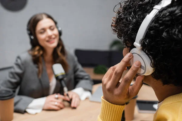 Jovem indiana homem ajustando fones de ouvido durante entrevista com host de rádio borrado em blazer cinza sorrindo perto de microfone e copo de papel no fundo borrado no estúdio de radiodifusão — Fotografia de Stock