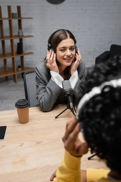 Happy brunette broadcaster adjusting headphones during interview with blurred indian man near professional microphone, coffee to go and smartphone with blank screen in modern radio studio — Stock Photo