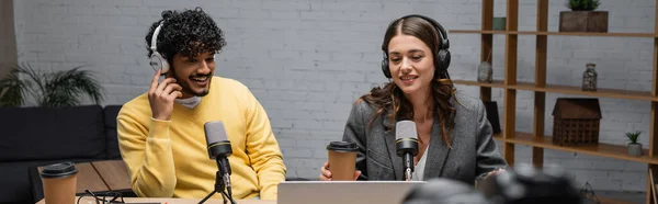 Cheerful interracial broadcasters in headphones, yellow jumper and grey blazer looking at laptop near microphones and coffee to go while working in modern radio studio, banner — Stock Photo