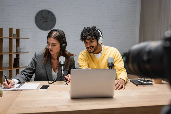 Brunette radio host in headphones writing in notebook close to positive indian colleague looking at laptop near professional microphones and smartphone with blank screen in studio — Stock Photo