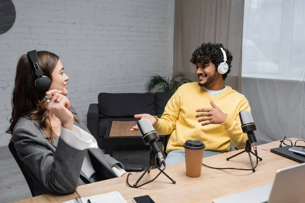 Happy multiethnic radio hosts in headphones talking near professional microphones, coffee to go, notebooks and smartphones on table in modern broadcasting studio with armchair on background — Stock Photo