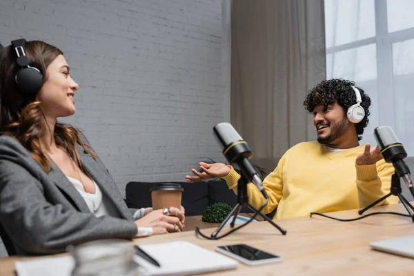 Joyful indian man in headphones and yellow jumper gesturing and talking to smiling colleague sitting with paper cup near microphones and blurred notebook with smartphone in broadcasting studio — Stock Photo