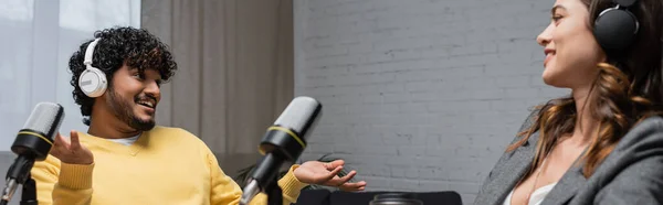 Кучерявий і веселий індійський чоловік в навушниках і жовтий джемпер розмовляє і жестикулює біля усміхненої брюнетки колеги і професійних мікрофонів в радіостудії, банер — стокове фото