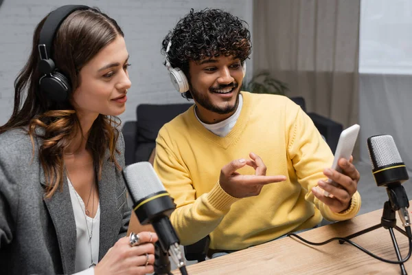 Sonriente podcaster indio en auriculares y jersey amarillo mostrando teléfono móvil a encantadora colega morena cerca de micrófonos profesionales en estudio de radiodifusión - foto de stock