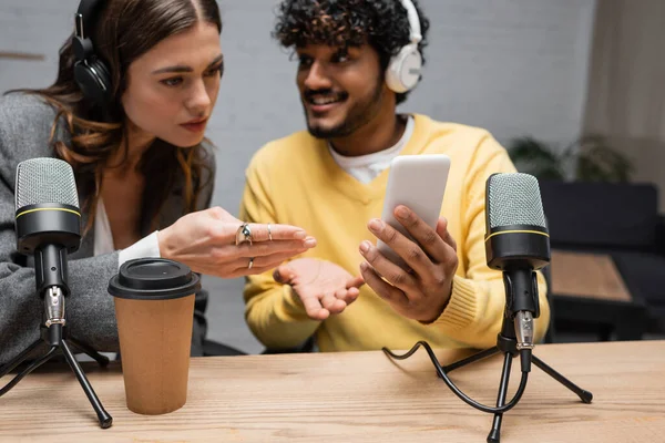 Serious radio host in headphones pointing at mobile phone in hands of positive indian colleague in yellow jumper near coffee to go and microphones in broadcasting studio — Stock Photo