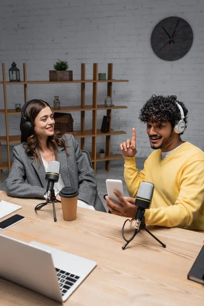 Cheerful and creative indian radio host showing idea gesture while looking at mobile phone near smiling colleague and professional microphones, laptop and smartphone with blank screen in radio studio — Stock Photo