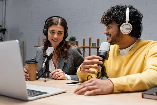 Smiling interracial broadcasters in headphones working with professional microphones near laptop, coffee to go and mobile phone with blank screen while recording podcast in radio studio — Stock Photo
