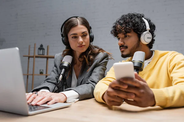 Charming brunette podcaster in headphones and grey blazer using laptop near young indian radio host holding blurred mobile phone while working in radio studio — Stock Photo