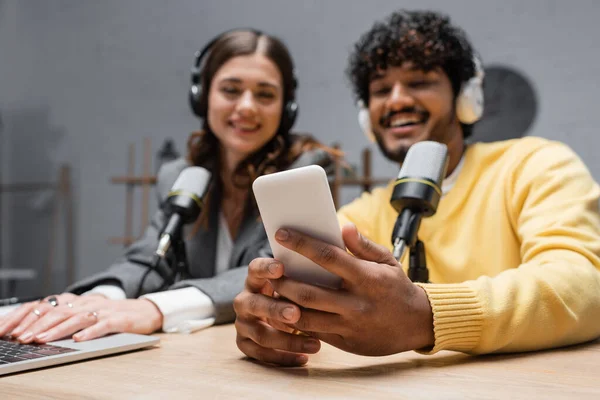 Cheerful indian radio host in headphones and yellow blazer holding smartphone near brunette colleague using laptop close to professional microphones in radio studio — Stock Photo