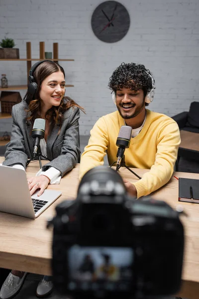 Homem indiano barbudo em fones de ouvido e jumper amarelo falando em microfone perto de notebook e colega sorridente usando laptop na frente de podcast de gravação de câmera digital em primeiro plano turvo — Fotografia de Stock
