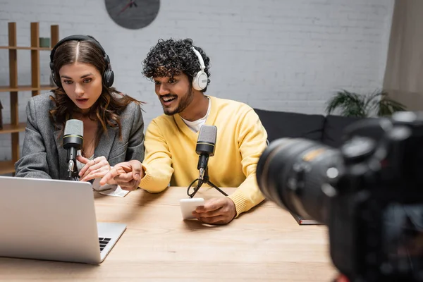 Homme indien joyeux dans les écouteurs et pullover jaune tenant smartphone et pointant vers un ordinateur portable près de collègue brune étonné et appareil photo numérique sur le premier plan flou en studio — Photo de stock