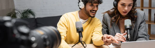 Cheerful indian podcaster in headphones and yellow jumper pointing at laptop near surprised colleague, microphones and professional digital camera on blurred foreground, banner — Stock Photo