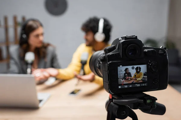 Screen of modern digital camera near blurred interracial colleagues in headphones recording podcast near laptop and mobile phone on table in radio studio — Stock Photo