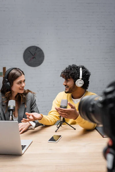 Positive indian podcaster in headphones and yellow jumper gesturing and pointing at colleague near laptop, microphones, mobile phone with blank screen and digital camera on blurred foreground — Stock Photo