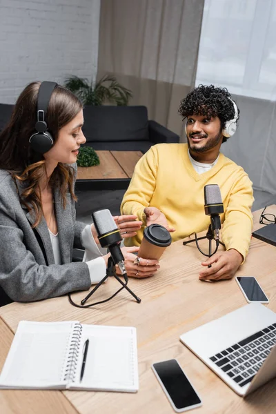 Cheerful indian man in headphones and brunette woman pointing at paper cup near professional microphones, laptop, notebooks and smartphones with blank screen in radio studio — Stock Photo