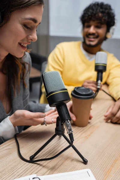 Щасливий подкастер вказує на виносний напій біля професійних мікрофонів та індійського колеги в жовтому джемпері, посміхаючись на розмитому фоні в радіостудії — стокове фото