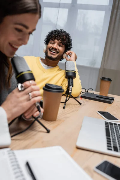 Hombre indio excitado en auriculares y jersey amarillo riendo cerca de colega borroso hablando en micrófono cerca de vasos de papel, cuadernos, computadora portátil y teléfono inteligente con pantalla en blanco en el estudio de radiodifusión - foto de stock