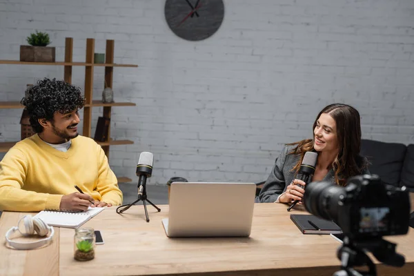 Smiling brunette announcer with microphone talking to young and curly indian colleague in yellow jumper writing in notebook near laptop, headphones and smartphone in modern radio studio — Stock Photo