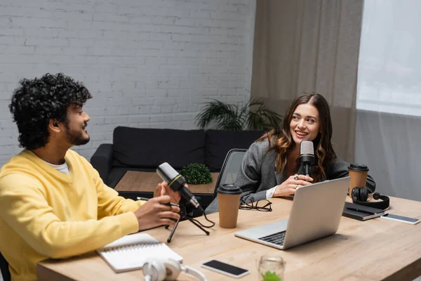 Cheerful brunette radio host looking at young indian colleague while talking near microphones, laptop, notebooks, paper cups and smartphones with blank screen in studio with couch on background — Stock Photo