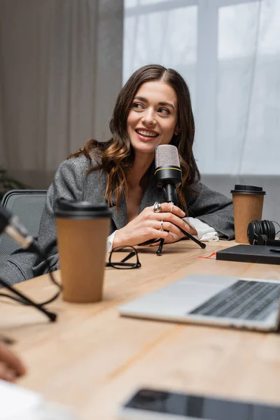 Happy and charming radio host looking away while talking in professional microphone near paper cups, blurred laptop and notebook on table in modern photo studio — Stock Photo