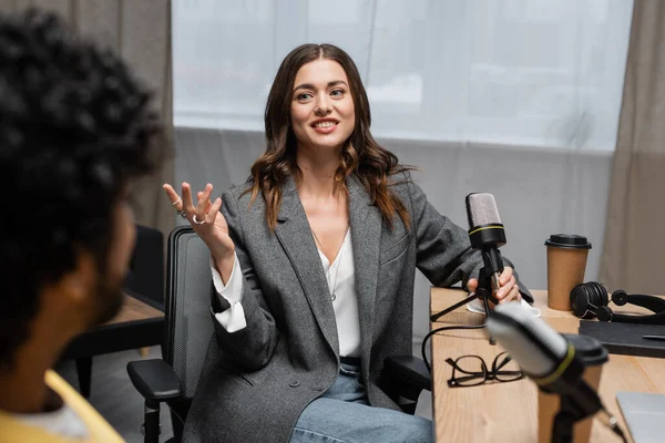 Pleased brunette podcaster in grey blazer holding microphone near headphones, notebook, paper cup and eyeglasses while gesturing and talking to indian man on blurred foreground in studio — Stock Photo