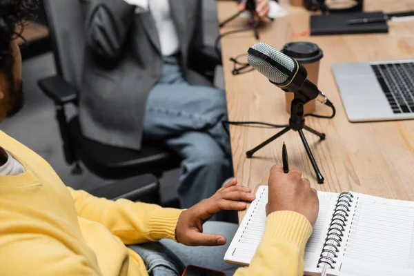 Обрезанный вид индийского вещателя, держащего ноутбук и ручку рядом с микрофоном, выпивку на вынос и ноутбук, сидя напротив коллеги на размытом фоне — стоковое фото