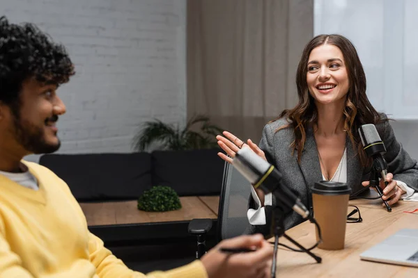 Excited brunette podcaster holding professional microphone and talking to blurred indian colleague in yellow jumper sitting near takeaway drink on table in radio studio — Stock Photo