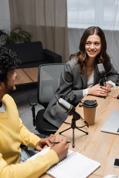 Charming brunette broadcaster in grey blazer holding professional microphone and smiling to blurred indian colleague sitting with pen and notebook near paper cup in radio studio — Stock Photo