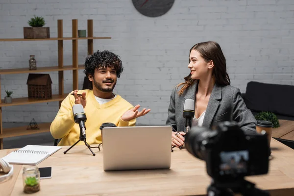Positive indian man in yellow jumper gesturing near microphones, laptop, notebook and smartphone while talking to smiling colleague sitting in blazer in front of blurred digital camera in studio — Stock Photo