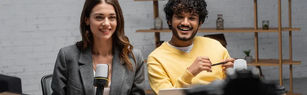 Cheerful brunette woman in grey blazer and curly indian man in yellow jumper smiling near professional microphones while working in modern radio studio, banner — Stock Photo