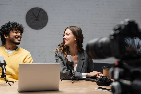 Cheerful radio host in grey blazer looking at young indian colleague in yellow jumper next to microphones, laptop, coffee to go, notebook and headphones in front of blurred digital camera in studio — Stock Photo