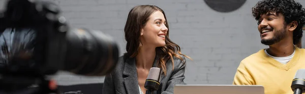 Bearded indian man in yellow jumper and charming brunette woman in grey blazer smiling at each other while recording podcast in front of studio microphones and blurred digital camera, banner — Stock Photo