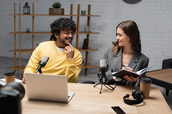 Smiling brunette woman in grey blazer writing in notebook close to young and cheerful indian man in yellow jumper near microphones, laptop, coffee to go, headphones and smartphone in studio — Stock Photo