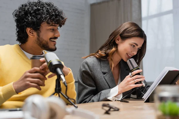 Young and curly indian man in yellow jumper smiling near charming colleague looking at notebook and talking in professional microphone in radio studio with blurred foreground — Stock Photo