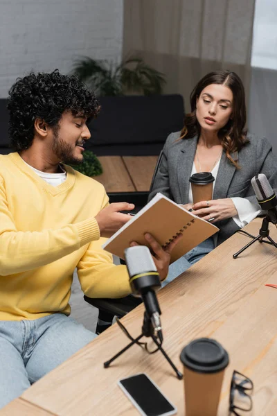 Homme indien barbu et bouclé montrant ordinateur portable à un collègue brune assis près de microphones professionnels, lunettes, gobelets de papier et téléphone portable avec écran blanc dans le studio de radiodiffusion — Photo de stock