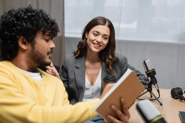 Curly indian radio host in yellow jumper showing notebook to happy brunette colleague in grey blazer sitting with coffee to go near professional microphones and headphones in radio studio — Stock Photo