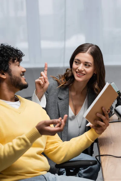 Joyful brunette woman in grey blazer showing idea gesture near curly and bearded indian colleague in yellow jumper sitting with notebook near table in radio studio — Stock Photo