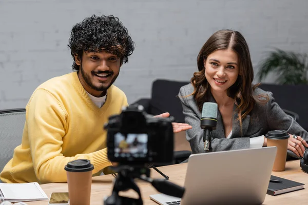 Cheerful indian man in yellow jumper pointing at happy brunette colleague near laptop, paper cup and notebooks while recording podcast on blurred digital camera in professional radio studio — Stock Photo