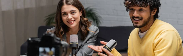Bearded and curly indian podcaster in yellow jumper talking in microphone near smiling brunette colleague wearing blazer in radio studio while recording podcast on blurred digital camera, banner — Stock Photo