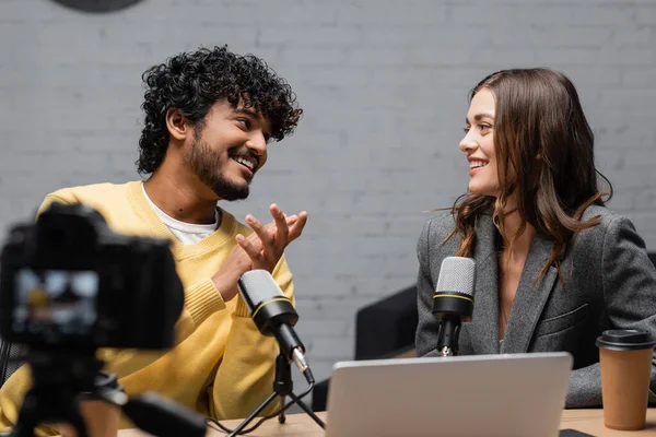 Curly and bearded indian man in yellow jumper smiling at charming colleague sitting in grey blazer near microphones, laptop and coffee to go in front of blurred digital camera in studio — Stock Photo