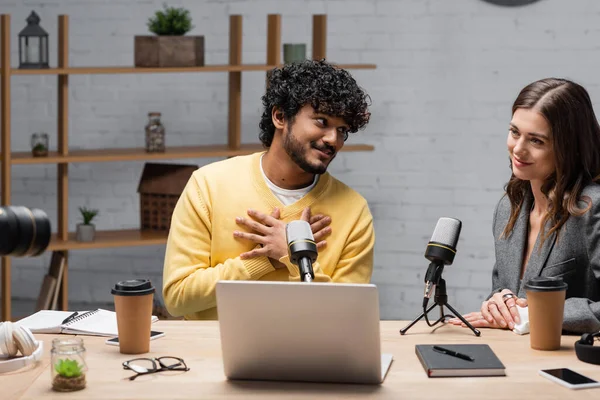 Honest indian podcaster touching chest and looking at smiling colleague near microphones, laptop, takeaway drinks, eyeglasses, notebooks and smartphones in broadcasting studio — Stock Photo
